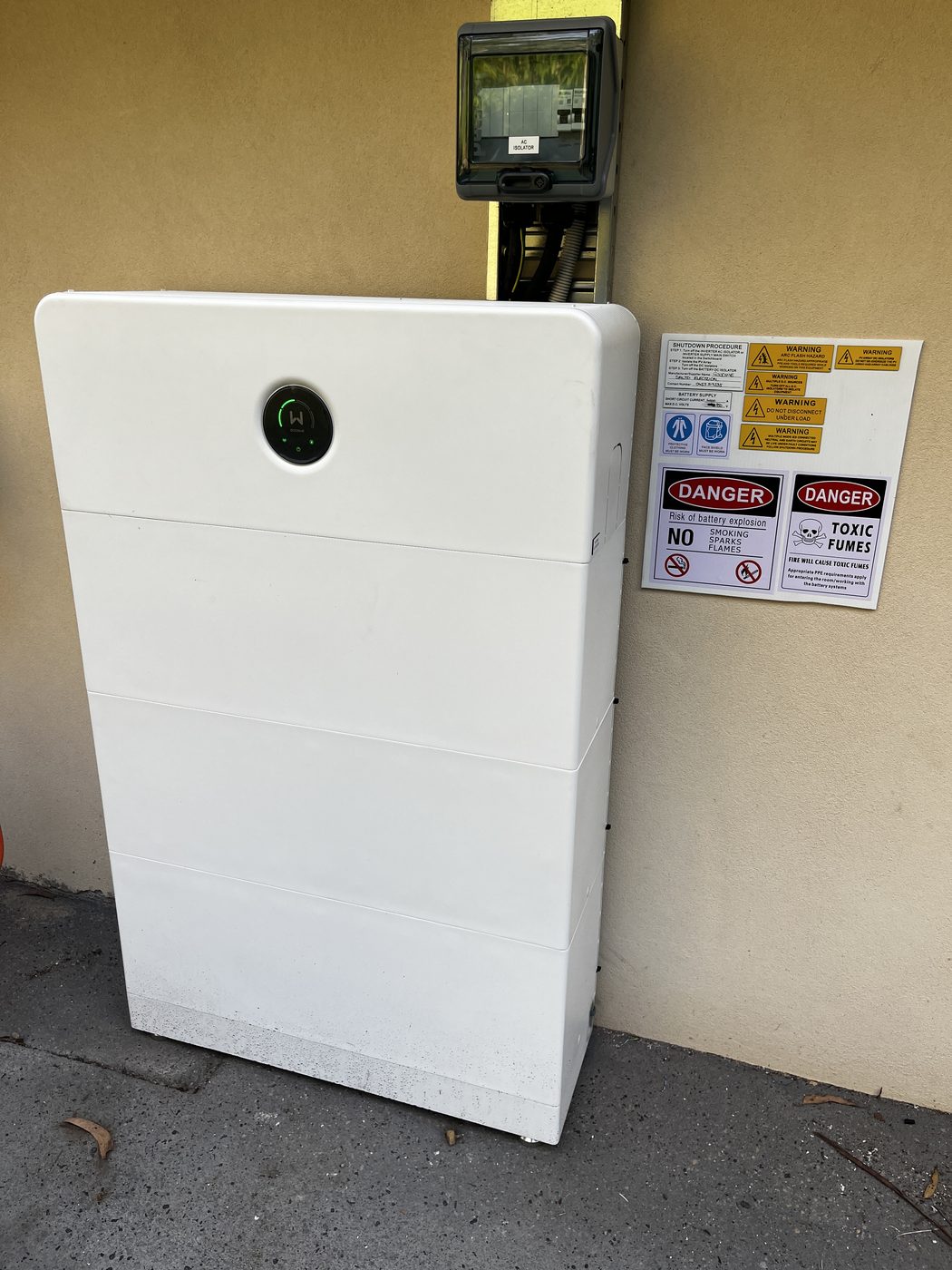 GoodWe stacked home battery installed outdoors against a rendered wall, powered up with its status ring lit, a labelled AC isolator in a weatherproof enclosure above it and the full shutdown procedure and hazard signage mounted alongside, installed by SALTD Electrical